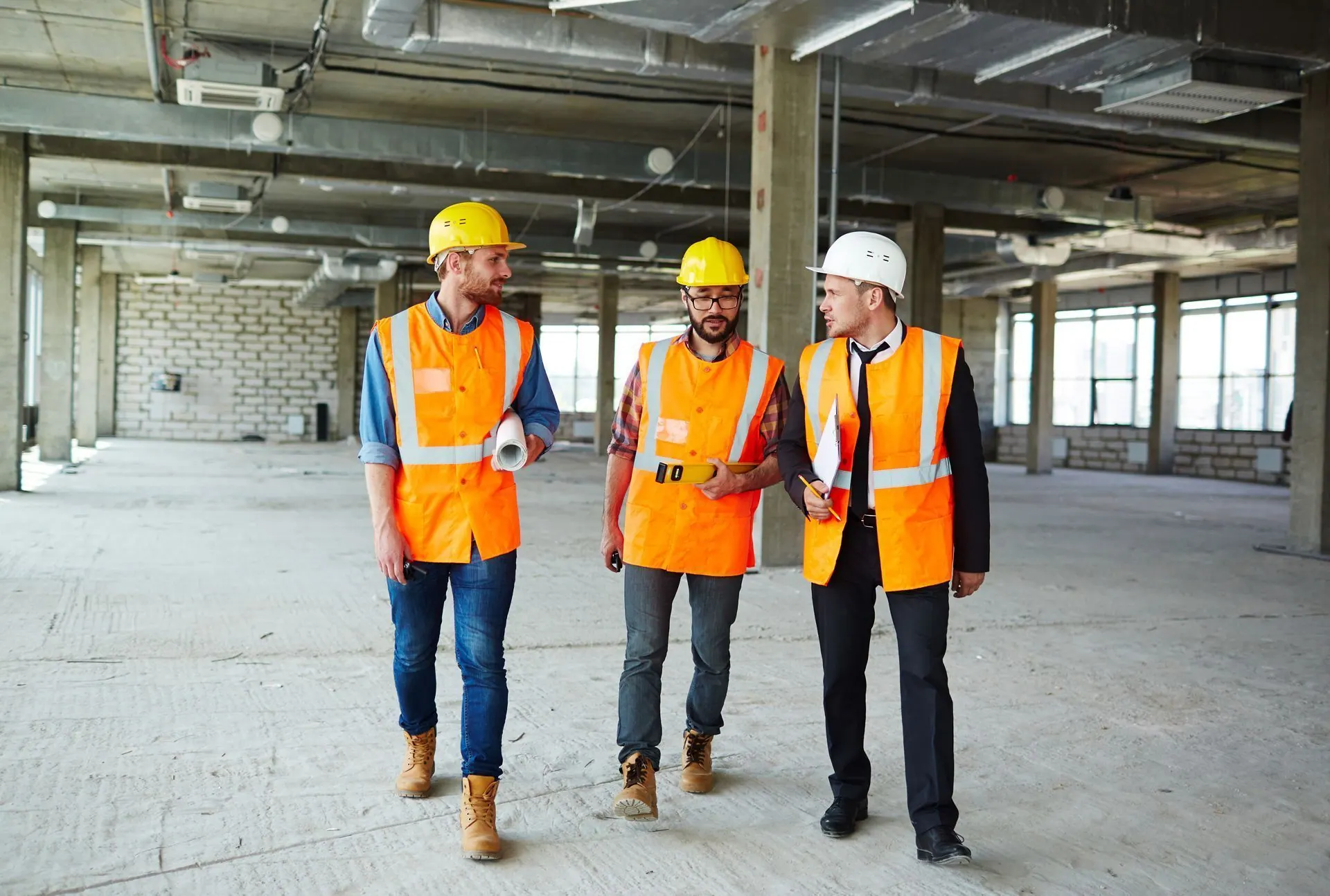 Three construction workers in hard hats and vests on a construction site; one holds blueprints.